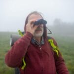 Starszy turysta z lornetką idzie we mgle po górskim szlaku